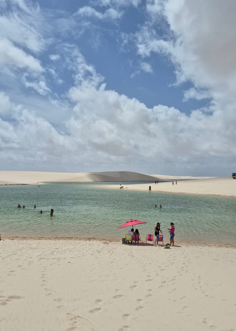 Turistas banhando em lagoa cristalina dos Lençóis Maranhenses com guarda-sol rosa e dunas ao fundo
