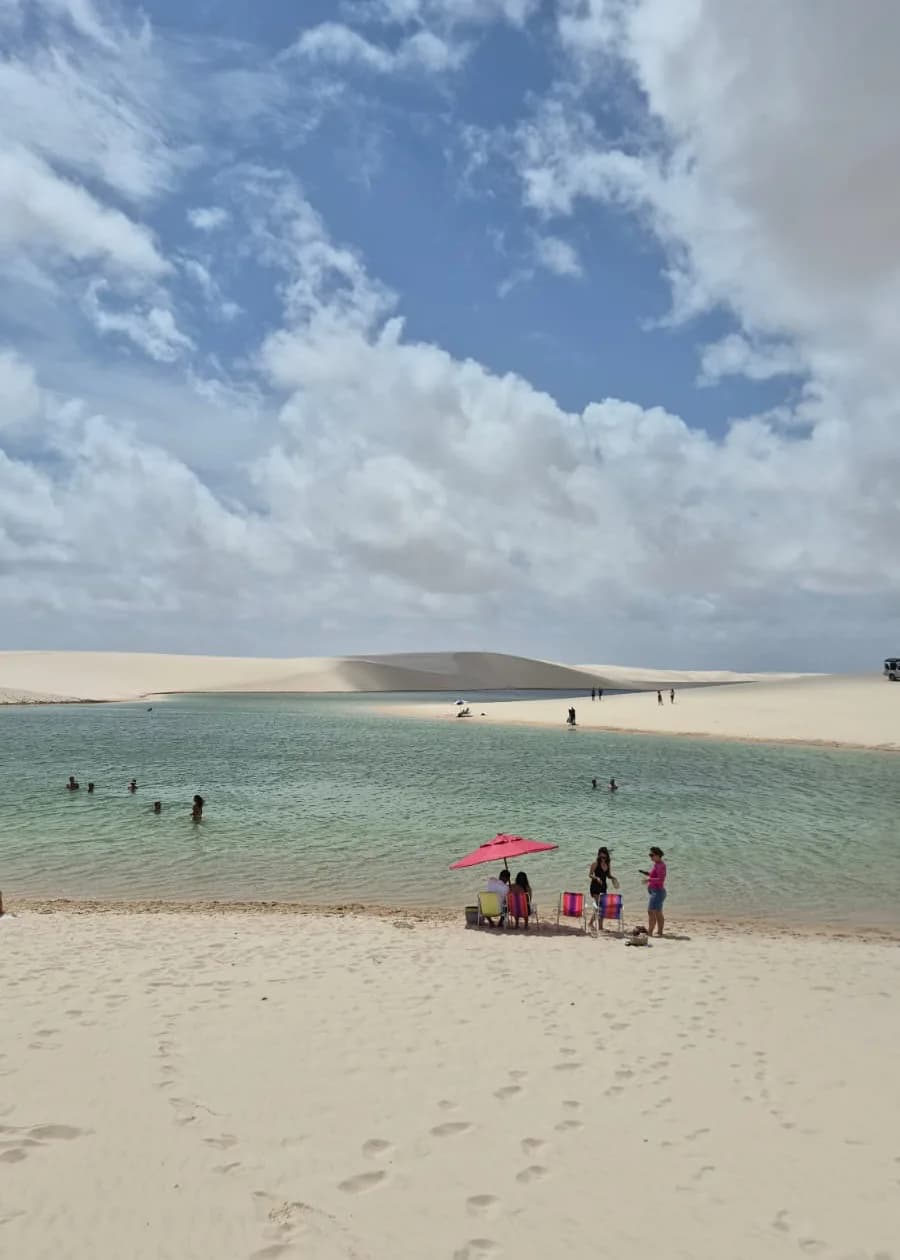 Turistas banhando em lagoa cristalina dos Lençóis Maranhenses com guarda-sol rosa e dunas ao fundo